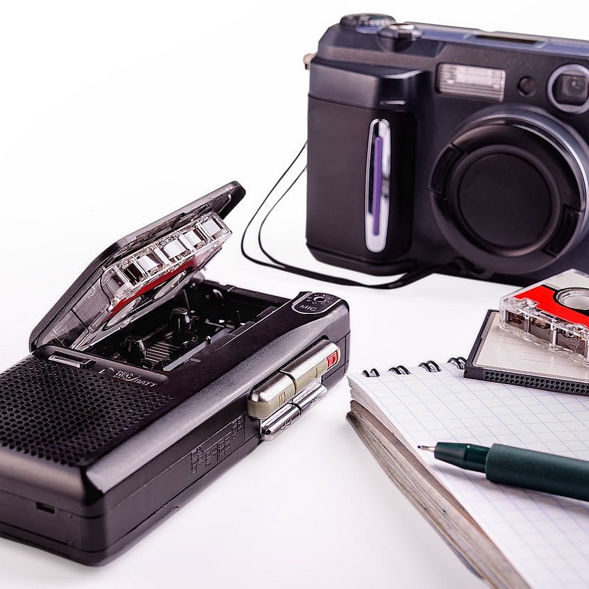 Das Bild zeigt eine Kamera, eine Diktiergerät sowie einen Block, einen Stift und eine Kassette.  