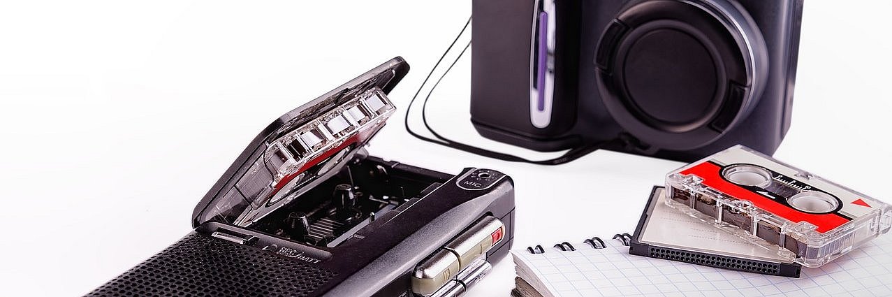 Das Bild zeigt eine Kamera, eine Diktiergerät sowie einen Block, einen Stift und eine Kassette.  