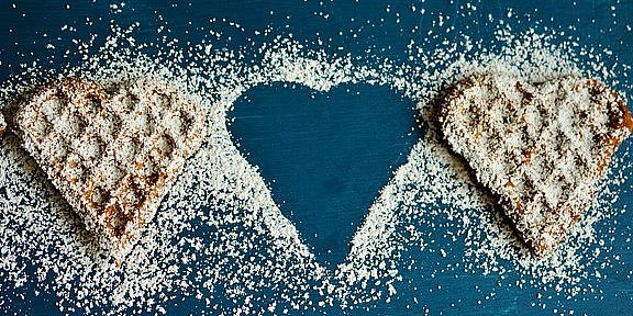 Auf einem blauen Untergrund liegen Waffelherzen, die mit Puderzucker bestreut sind. Von rechts sind eineinhalb Waffelherzen zu sehen. Eine Waffelherz wurde ausgelassen. Dies ist am Puderzuckerrand zu sehen, der das Herz andeutet. Daneben ist eine weiteres Waffelherz angelegt. 