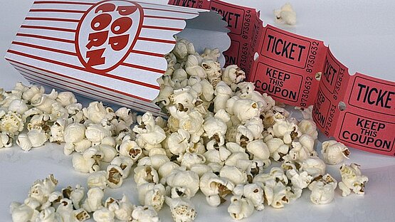 Das Bild zeigt links eine umliegende Popcorntüte. Die Popcorntüte ist weiß und hat rote Streifen. Ein Oval in der Mitte der Tüte trägt den roten, fetten Schriftzug "Pop Corn". Die oberen Kanten der Tüte sind in Wellen abgerundet. Aus der Tüte heraus ist Popcorn gefallen, welches rechts neben und vor der Tüte liegt. Hinter der Tüte stehen im Zick-Zack sechs rote Kino-Eintrittskarten. Die Tickets sind rechteckig. Auf diesen steht in schwarzer Schrift und jeweils eingerahmt, untereinander "Ticket" und "Keep this coupon". Rechts neben den beiden Schriftzügen steht untereinander zweimal die Ticketnummer.